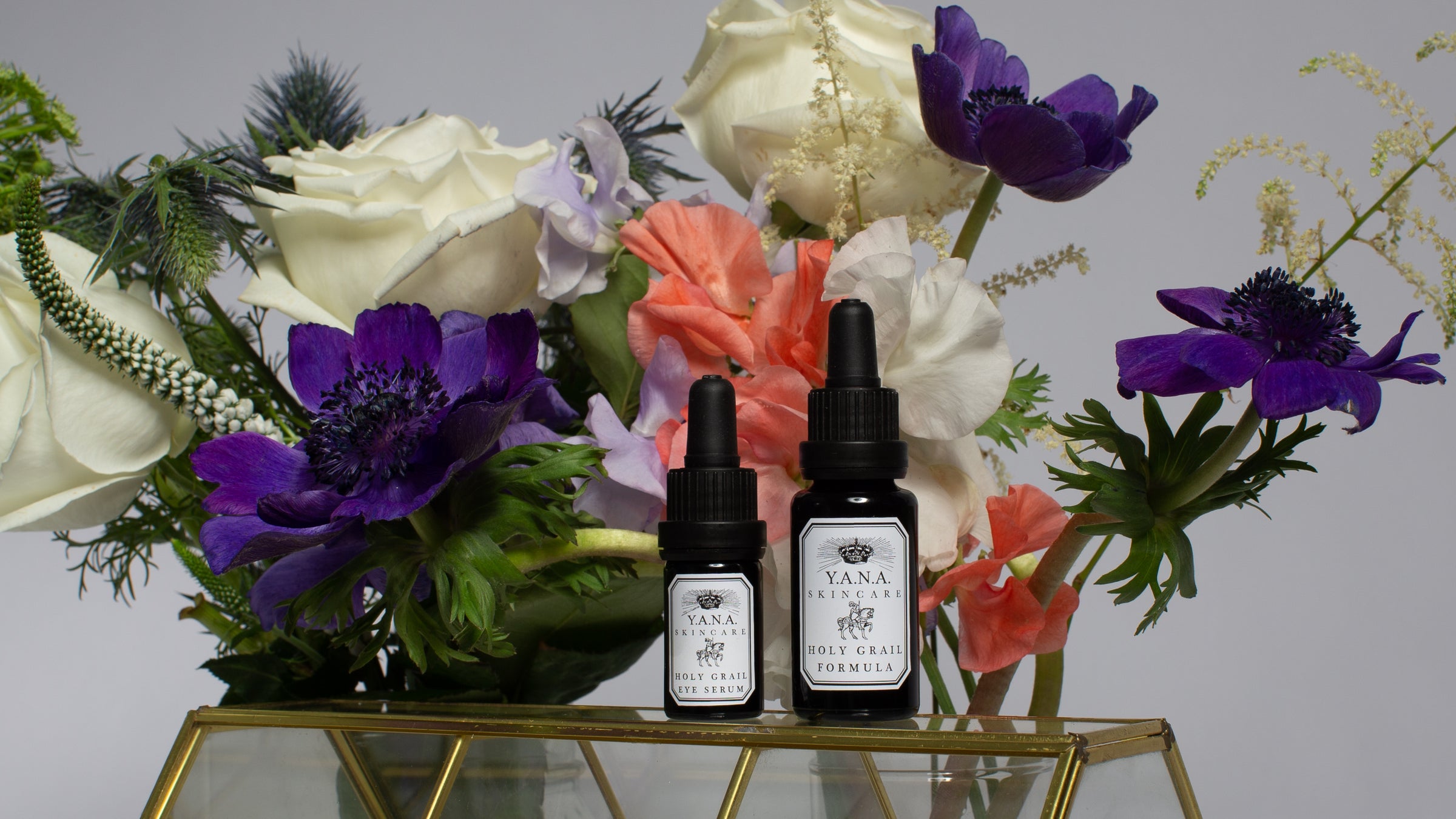 Two Yana Skincare Holy Grail Serum bottles on a glass cabinet surrounded by white roses, purple poppies, and other various flowers.