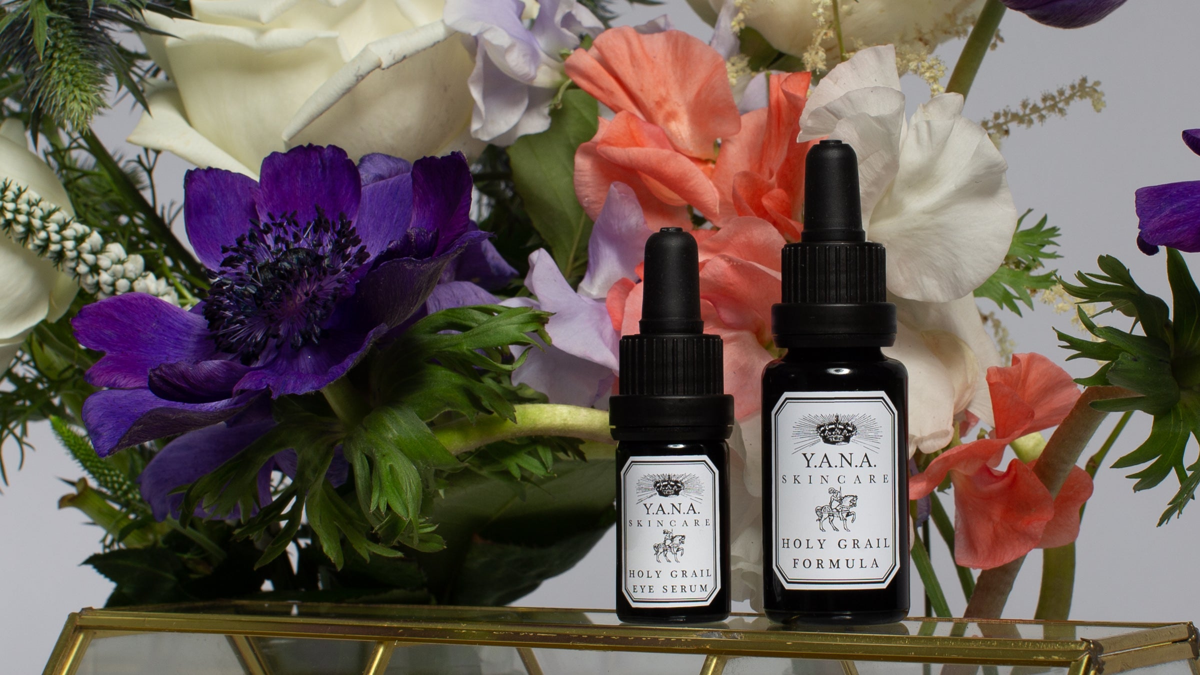 Two Yana Skincare Holy Grail Serum bottles on a glass cabinet surrounded by white roses, purple poppies, and other various flowers.