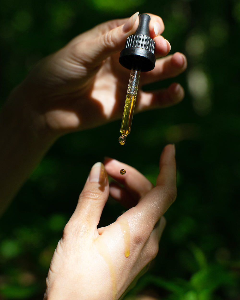 Hand dripping YANA SKINCARE Holy Grail Serum onto another hand, in a forest setting.