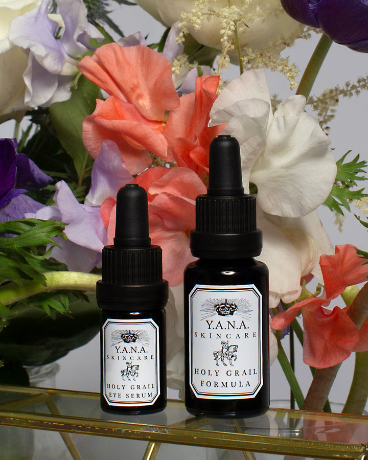 Yana Skincare Face and Eye Serum bottles on a glass case in front of various colored exotic flowers.